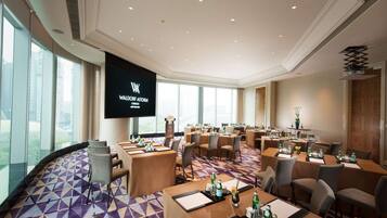 Un salón de conferencias con varias mesas y sillas, una pantalla grande mostrando 'WALDORF ASTORIA CHICAGO', y ventanas amplias con vistas a la ciudad.