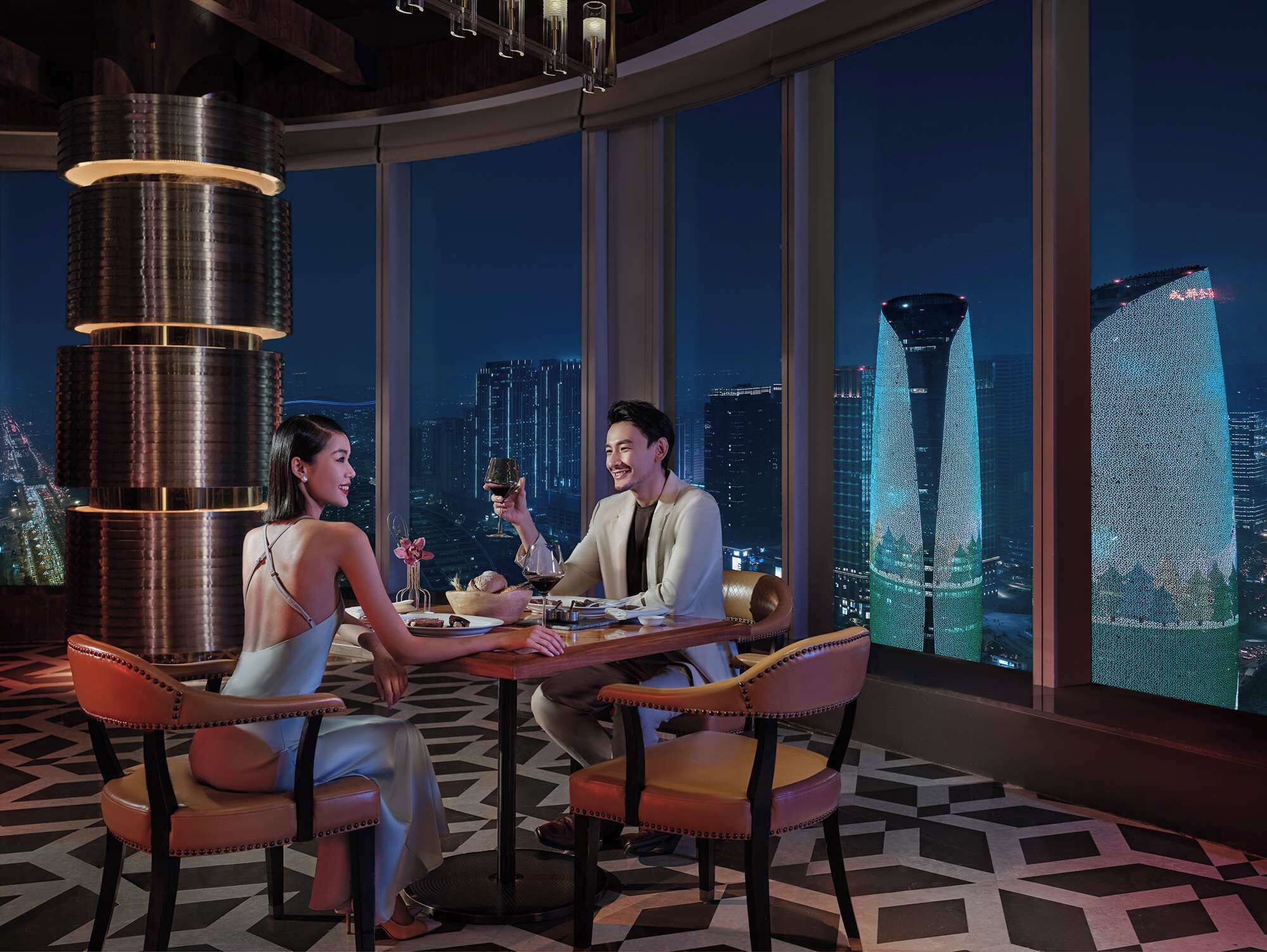 A high-rise hotel room with a panoramic city view, a modern chandelier, and a dining table set for two.