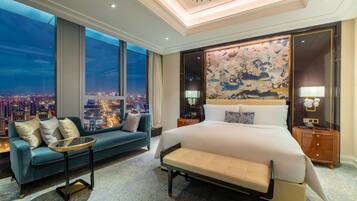 Una habitación de hotel moderna con una cama grande, un sofá, una mesita y vistas a un paisaje urbano nocturno.