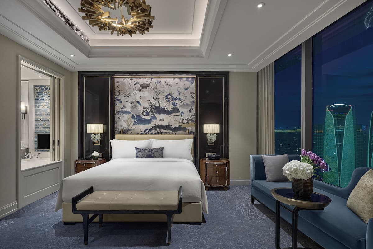 A modern hotel room with a large bed, a bench, a sofa, and a view of a cityscape.