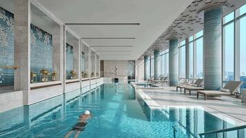 A modern indoor swimming pool with clear water, a swimmer, and lounge areas with views of the city.