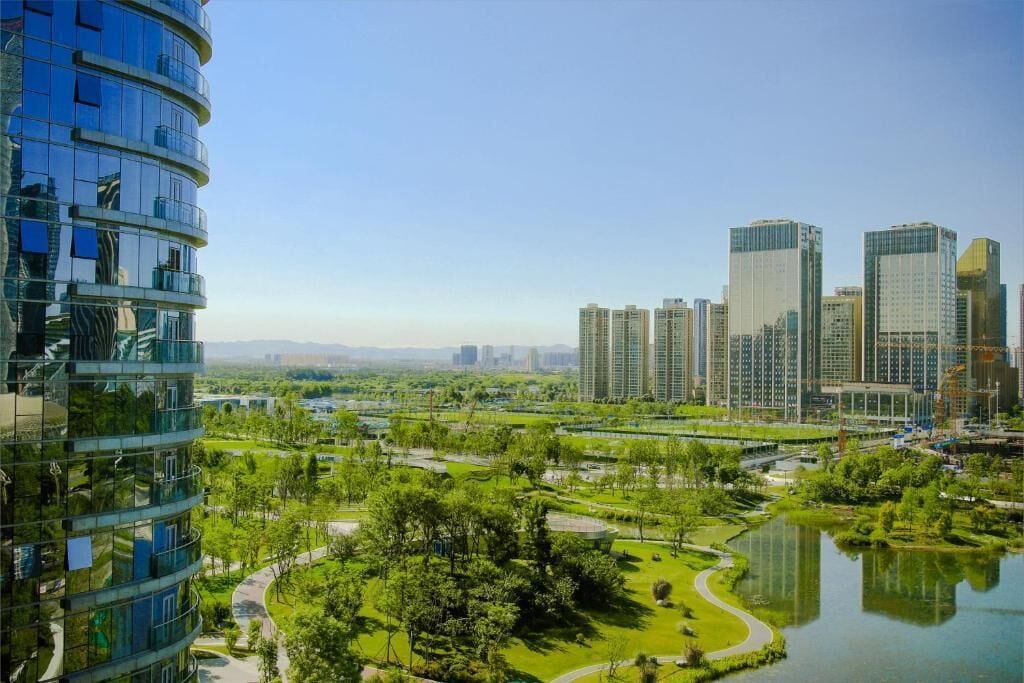 A modern high-rise building with a curved glass facade, surrounded by a park with a lake and walking paths.