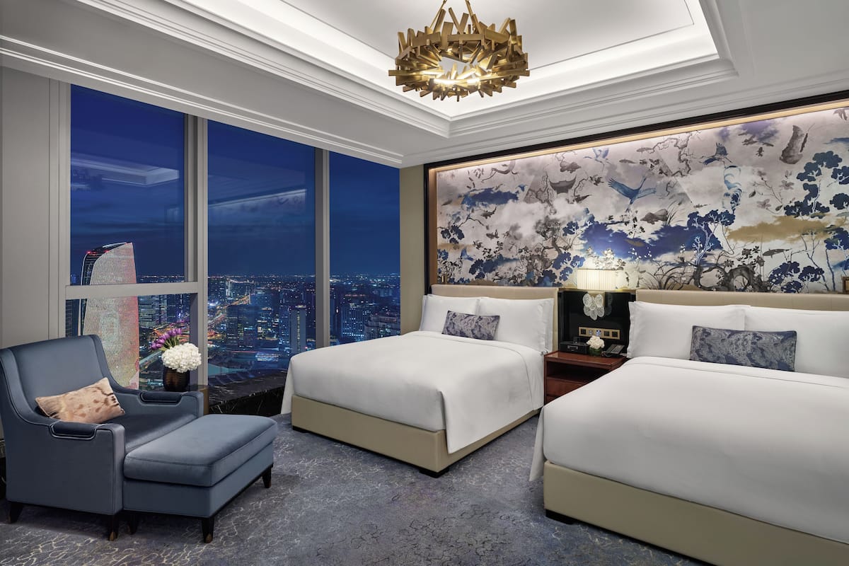 A hotel room with a city view, two beds, a chair, a chandelier, and a decorative wall.