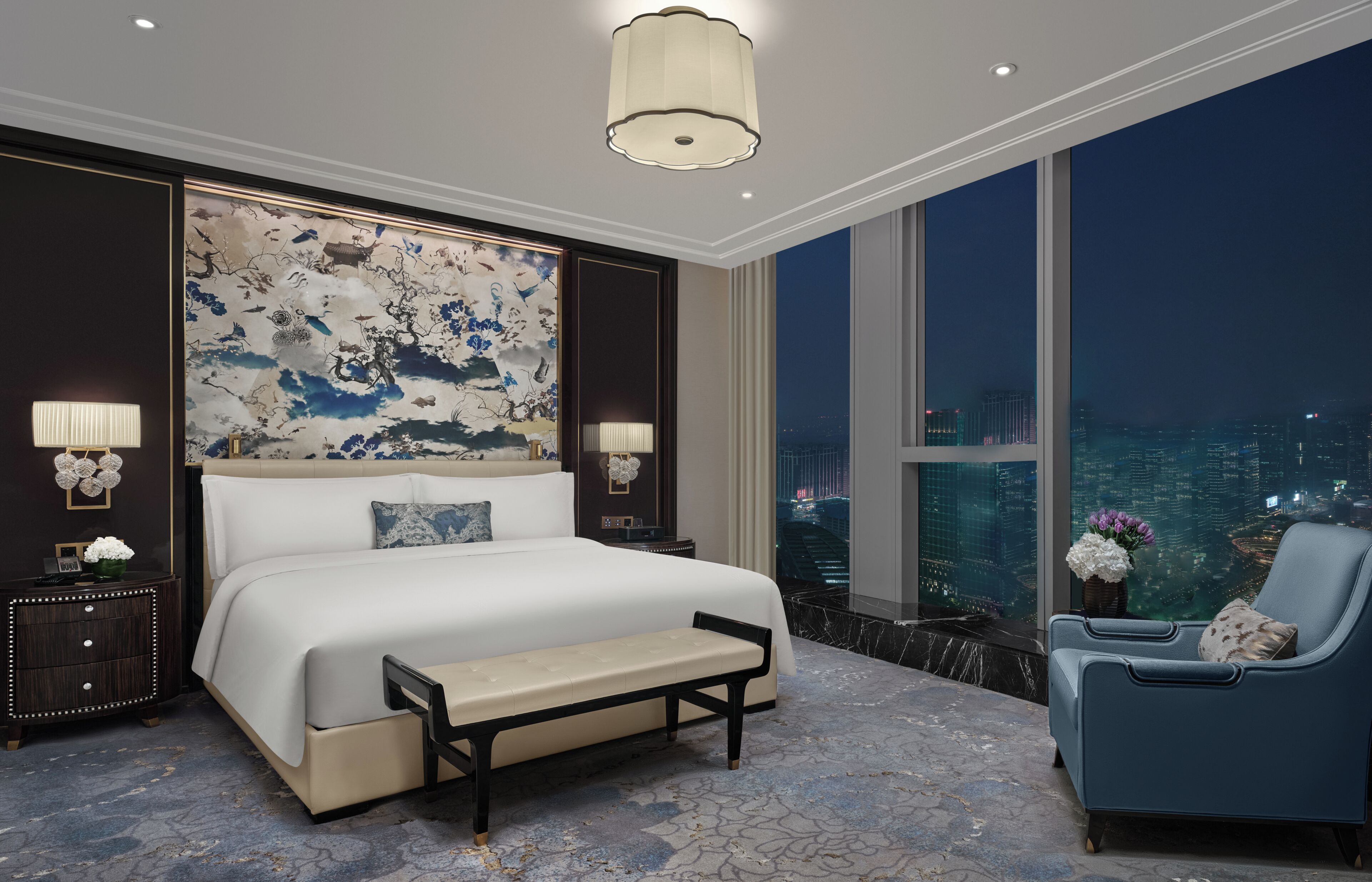 Een moderne slaapkamer met een groot bed, nachtlampjes, een bankje, een stoel en uitzicht over de stad.