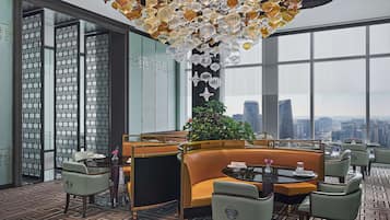 A modern dining area with a city view, featuring a patterned floor, a central seating area with a sofa and chairs, and a decorative ceiling light fixture.