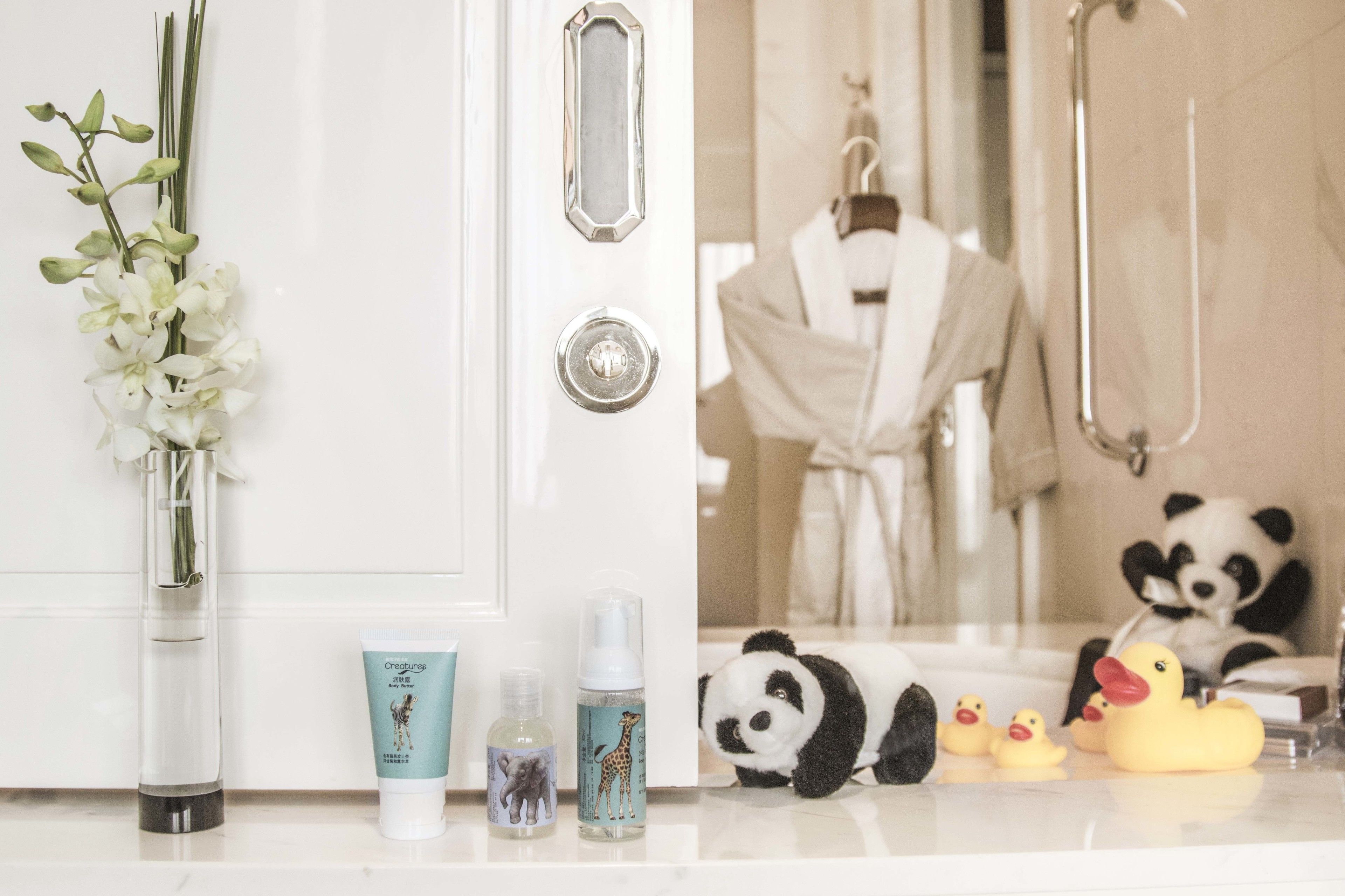 A bathroom with a white door, a vase with white flowers, a bottle of lotion, and plush toys including a panda and rubber ducks.