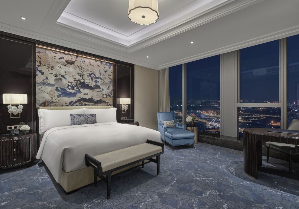 Habitación de hotel con una cama grande, un escritorio, una silla, una mesita de noche y un ventanal con vistas a la ciudad.