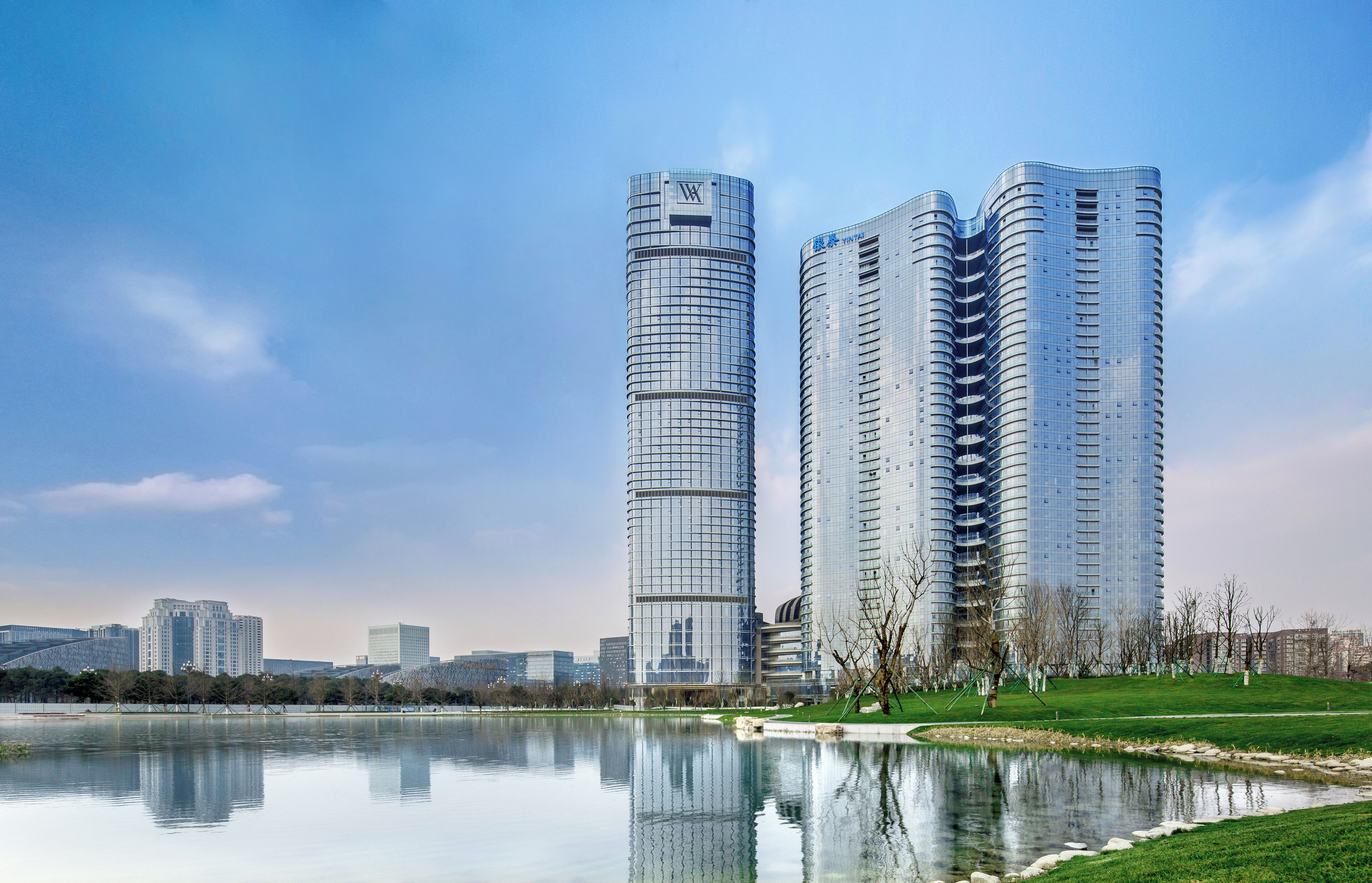 Two modern high-rise buildings with reflective glass facades, a calm lake, and a park area with trees and a pathway.