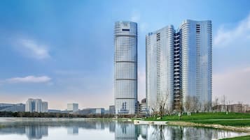 Two modern high-rise buildings with reflective glass facades, a calm lake, and a park area with trees and a pathway.