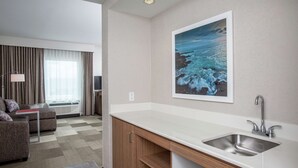 A modern hotel room with a kitchenette, a sofa, a large window with curtains, and a painting of a seascape. - Hampton Inn & Suites Oahu/Kapolei (Kapolei)