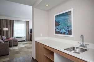 Una habitación de hotel moderna con kitchenette, un sofá, un ventanal con cortinas y un cuadro de paisaje marino.
