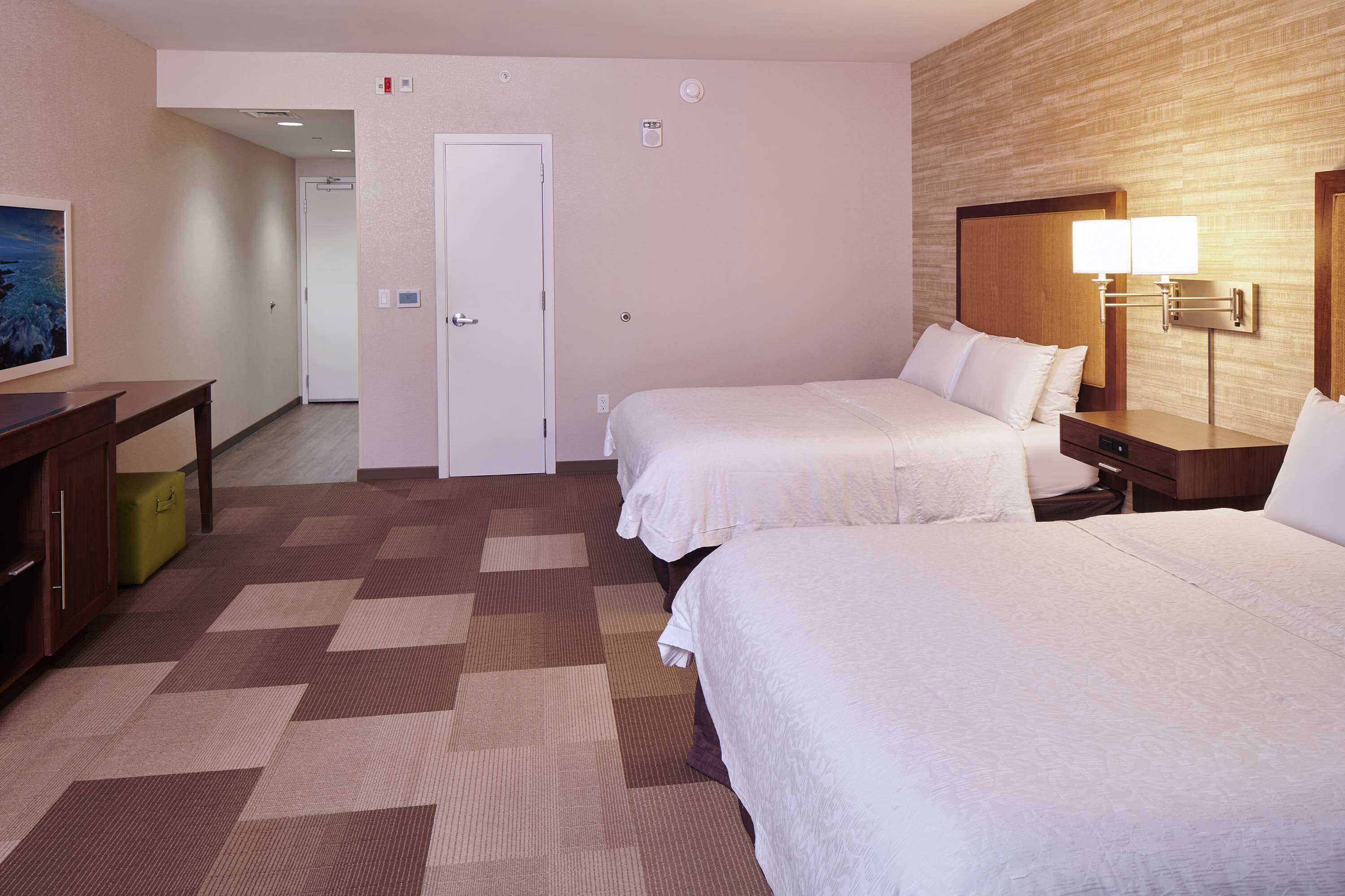 A hotel room with two beds, a desk, a lamp, and a door.