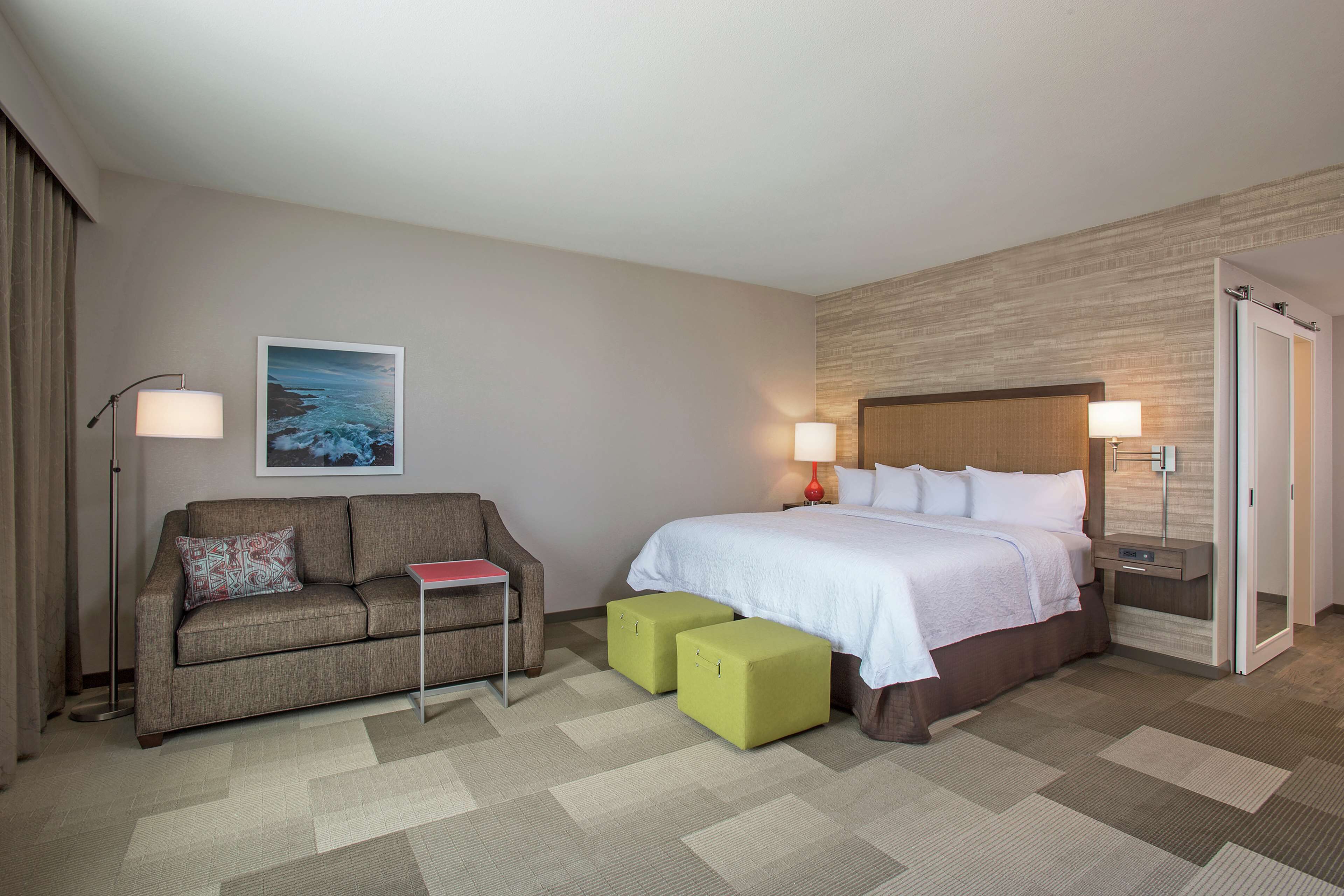 A hotel room with a bed, a sofa, two ottomans, a nightstand, a lamp, and a framed picture on the wall.
