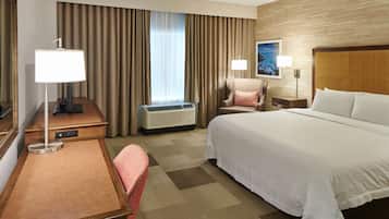 Ein Hotelzimmer mit Bett, Schreibtisch, Stuhl, Lampe, Klimaanlage und einem fensterbepflanzten Fenster.