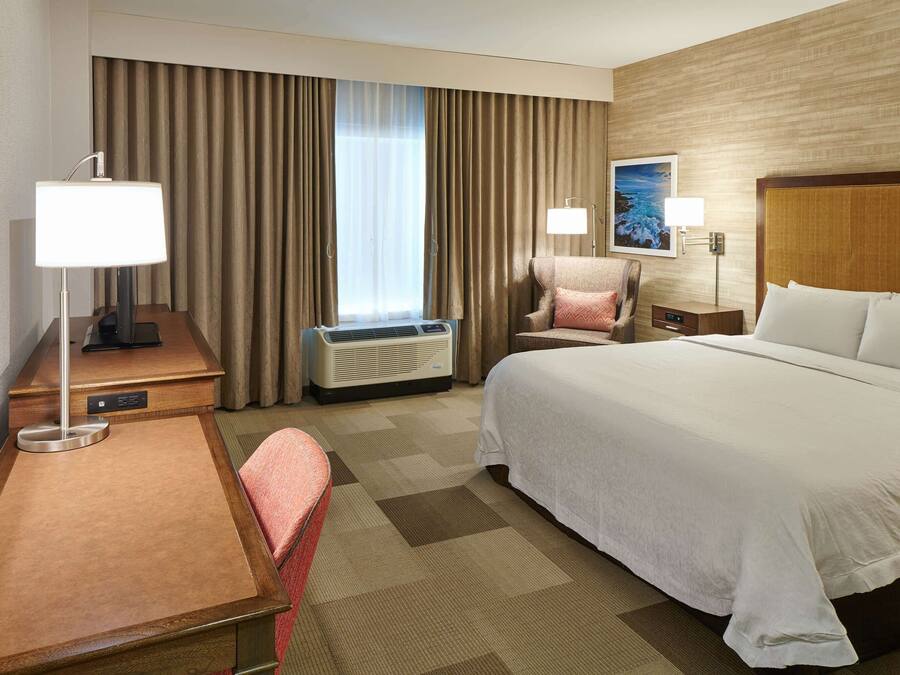 Ein Hotelzimmer mit Bett, Schreibtisch, Stuhl, Lampe, Klimaanlage und einem fensterbepflanzten Fenster.