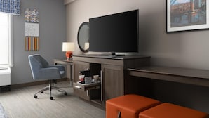 A hotel room with a large flat-screen TV, a desk with a lamp, a chair, and a sofa. There are artworks on the wall and a window with blinds. - Hampton Inn & Suites Yonkers - Westchester (Yonkers)
