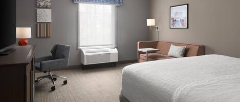 A hotel room with a large bed, a desk with a lamp, a chair, a window with blinds, and a wall with artwork.