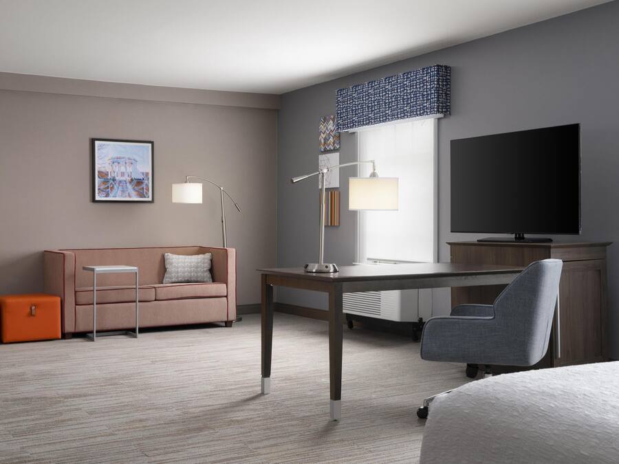 A hotel room with a sofa, a desk, a chair, a TV, and a lamp.