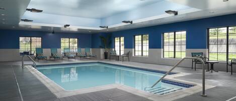 An indoor swimming pool with clear blue water, surrounded by lounge chairs and tables, and large windows offering views of the outdoors.