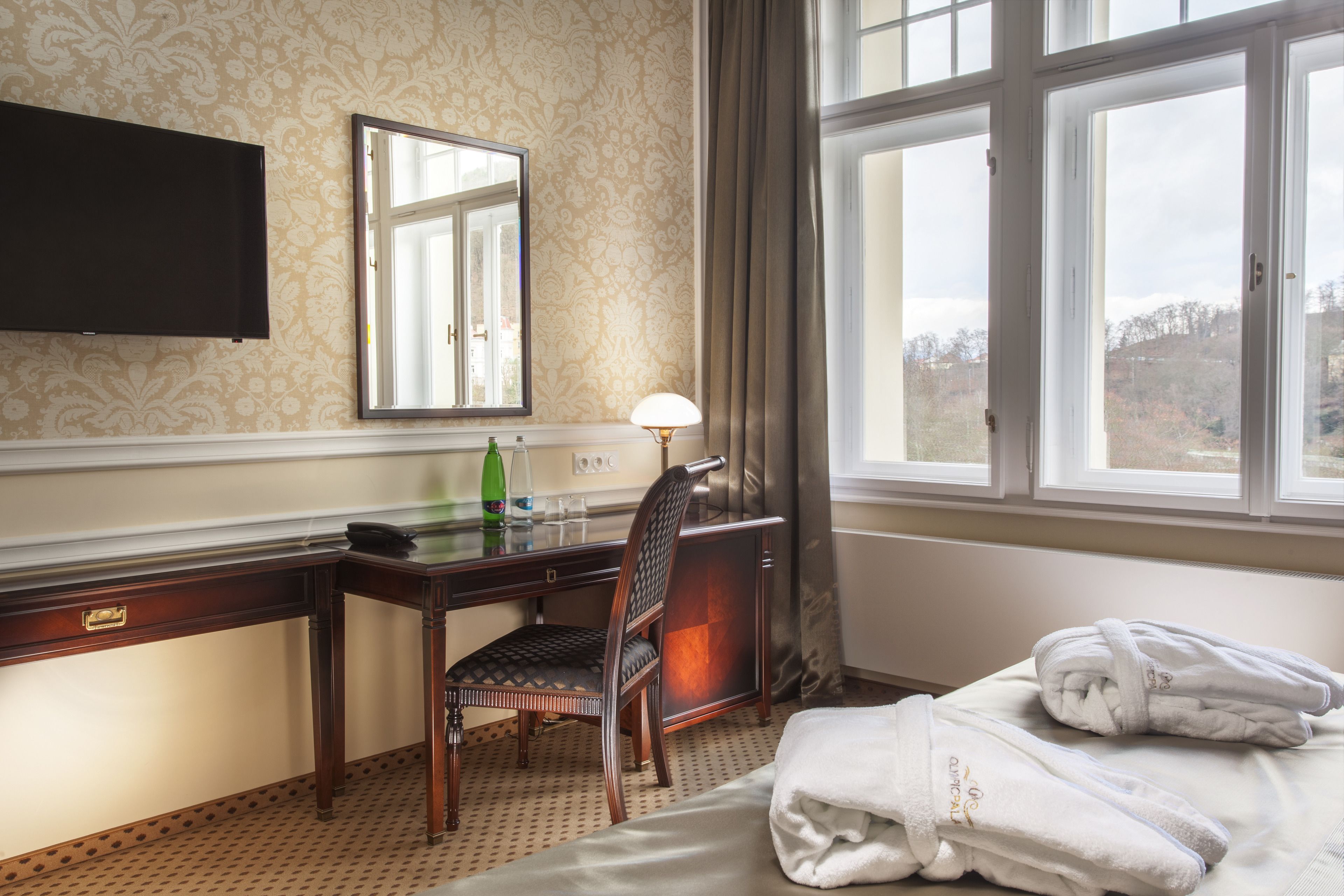 Ein Hotelzimmer mit Bett, Schreibtisch mit Stuhl, Flachbildfernseher, Fenster mit Vorhängen und Blick ins Freie.
