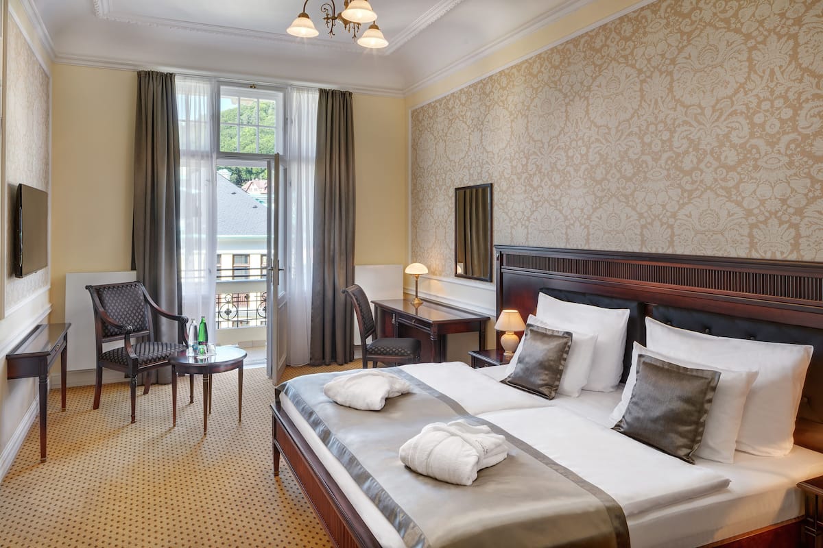 A hotel room with a large bed, two chairs, a small table, a desk, a lamp, and a large window with curtains.