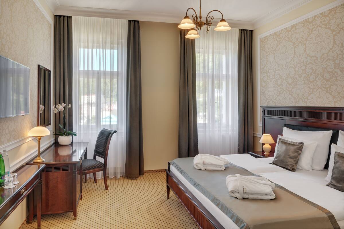 A hotel room with a bed, bedside table, lamp, chair, and a large window with curtains.