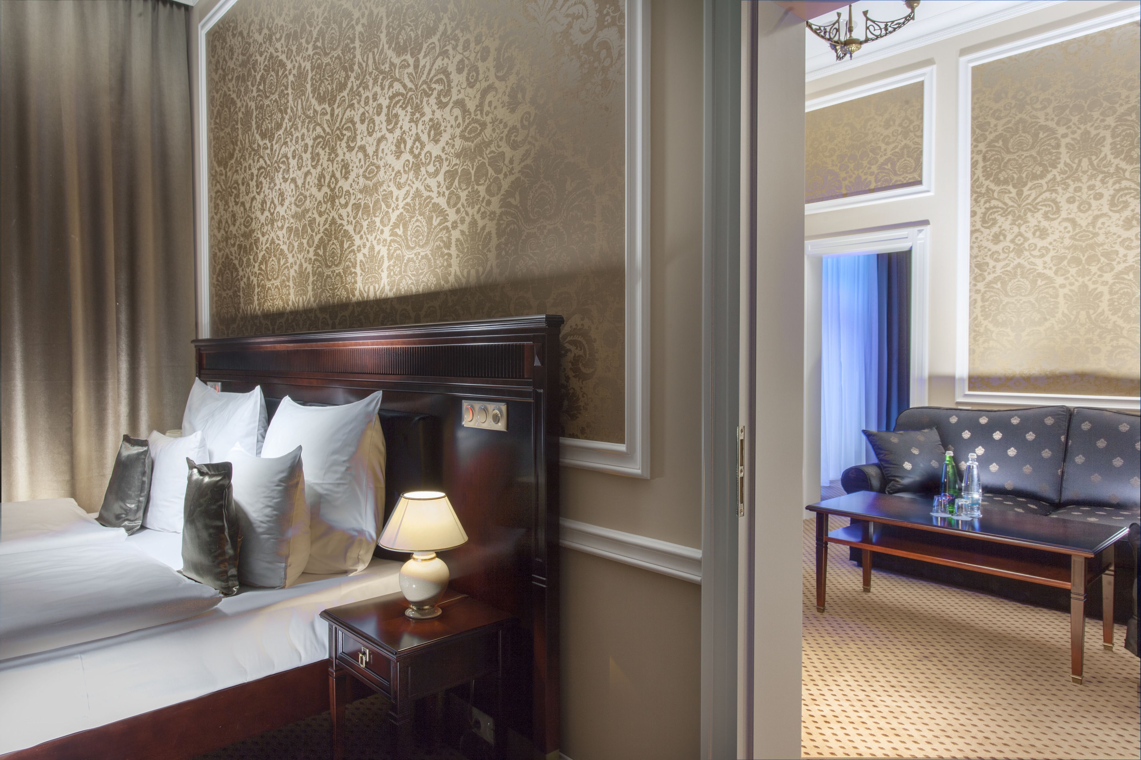 A hotel room with a bed, nightstand, lamp, and a sofa in the adjacent living area.