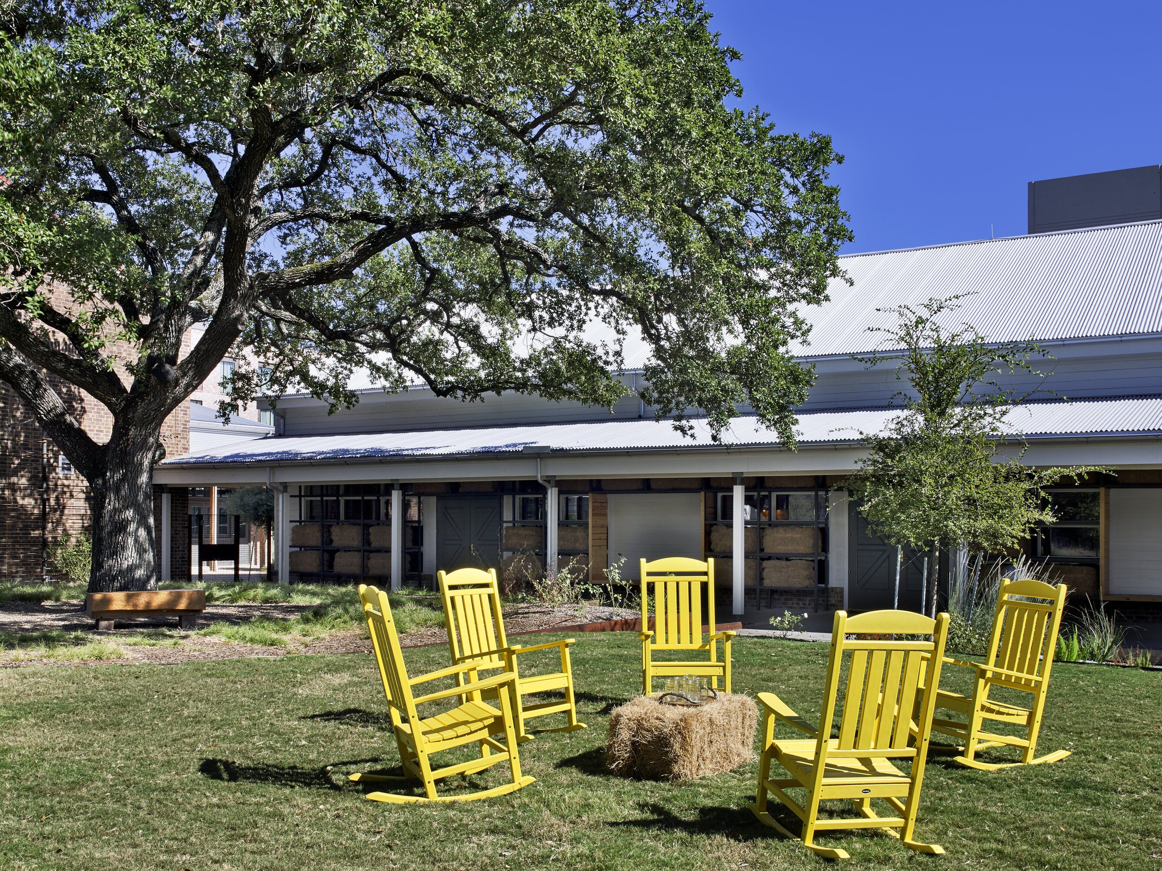 A building with a covered patio area, outdoor seating with yellow chairs, and a large tree providing shade.