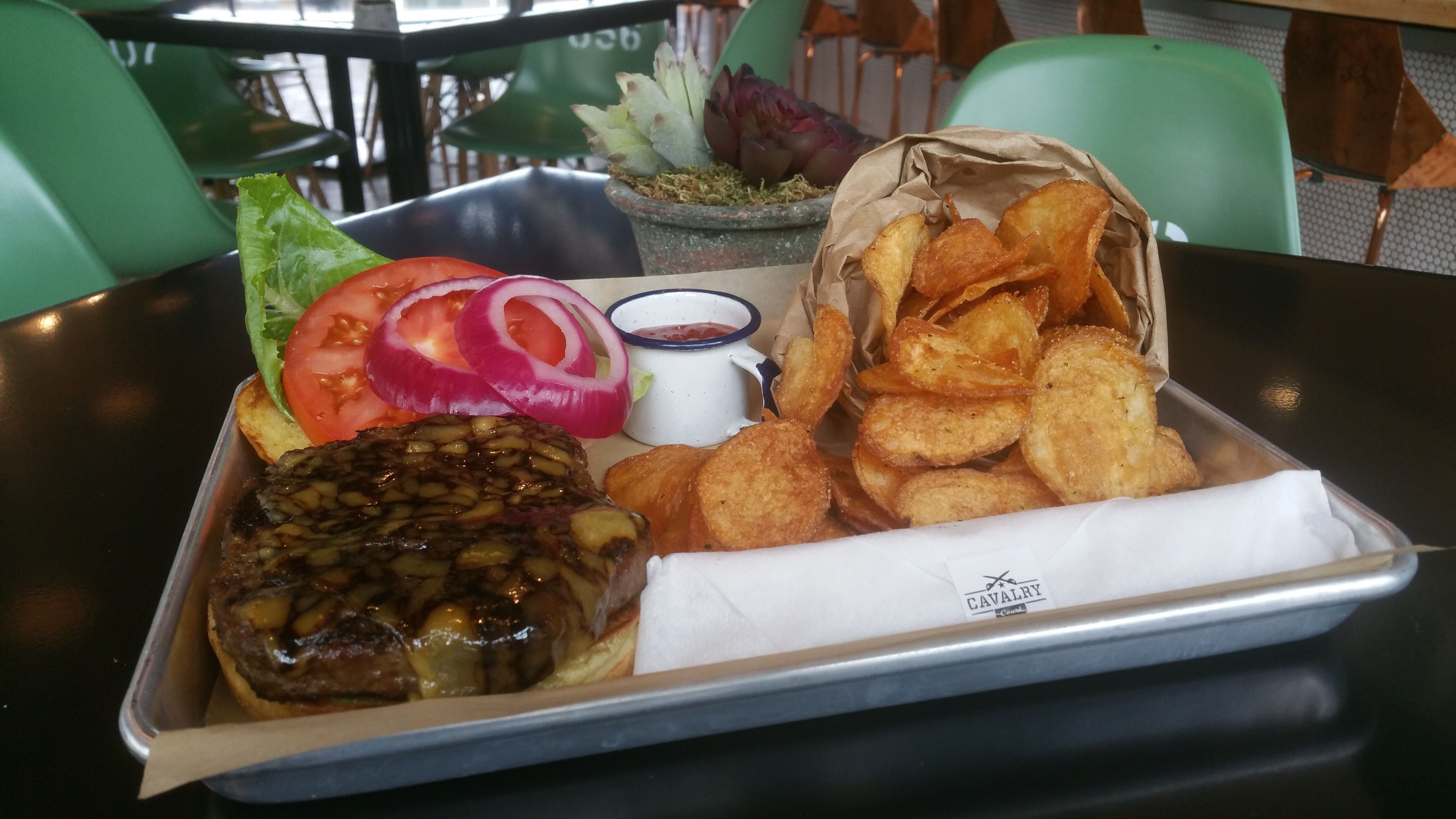 A tray with a meat dish, potato wedges, sliced tomatoes, red onions, and a small cup of sauce on a table.