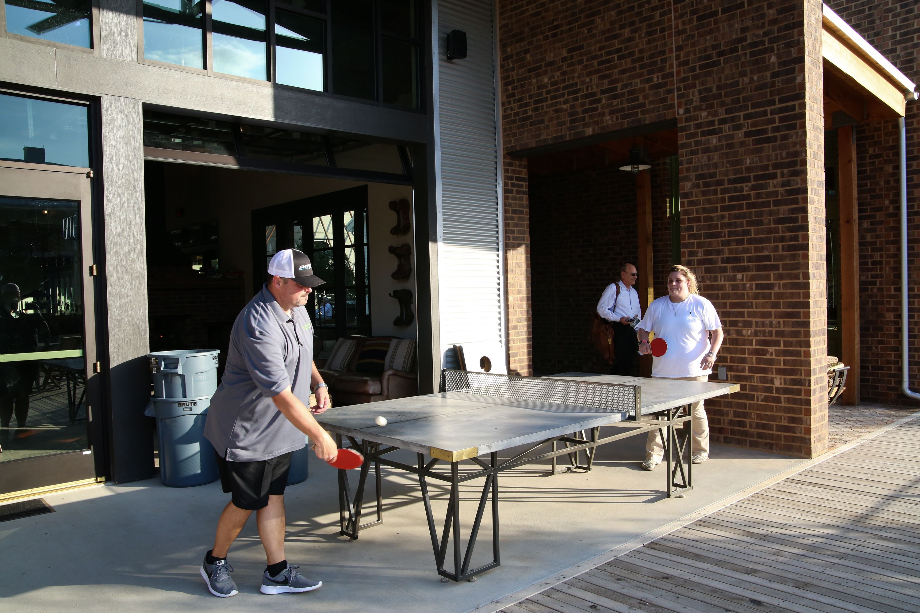 An outdoor ping pong table set up on a wooden deck in front of a building with a brick facade.