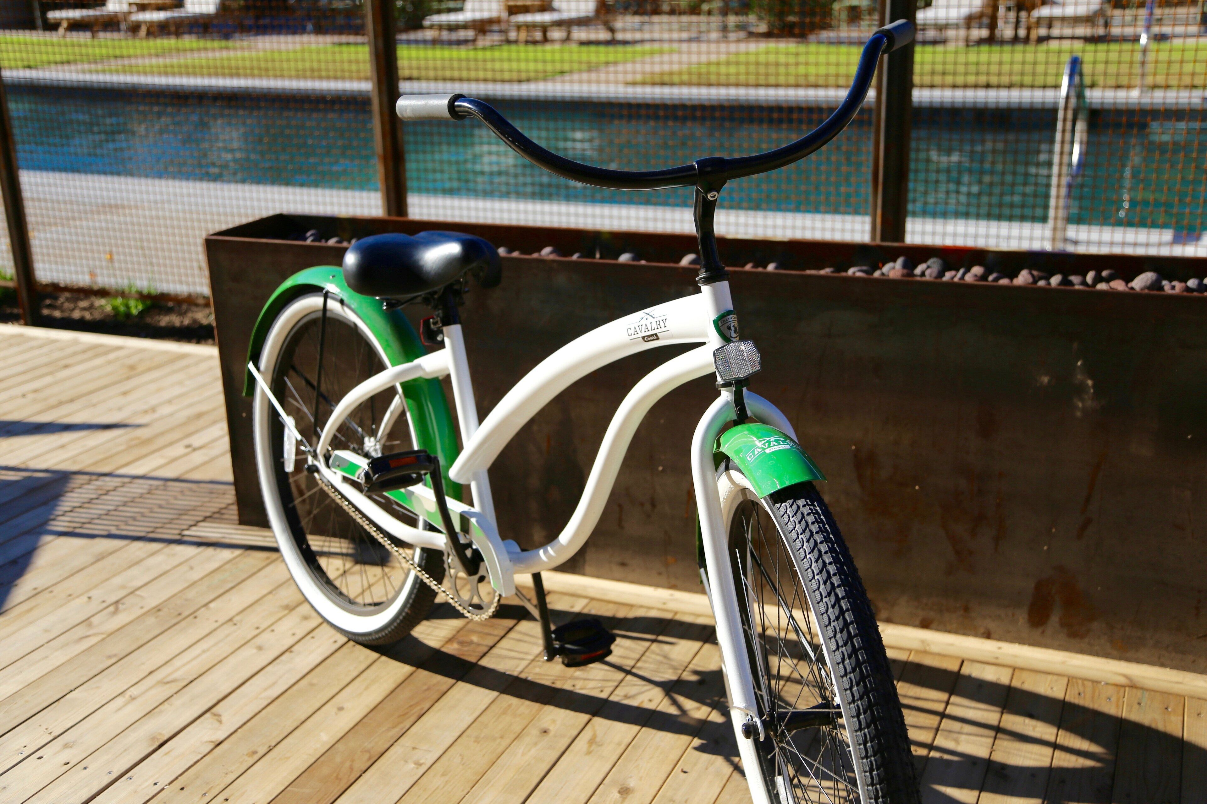 Uma bicicleta ergométrica com assento verde e estrutura branca, posicionada sobre um deck de madeira com guarda-corpo de metal e uma piscina ao fundo.