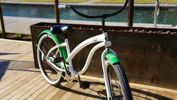 Uma bicicleta ergométrica com assento verde e estrutura branca, posicionada sobre um deck de madeira com guarda-corpo de metal e uma piscina ao fundo.