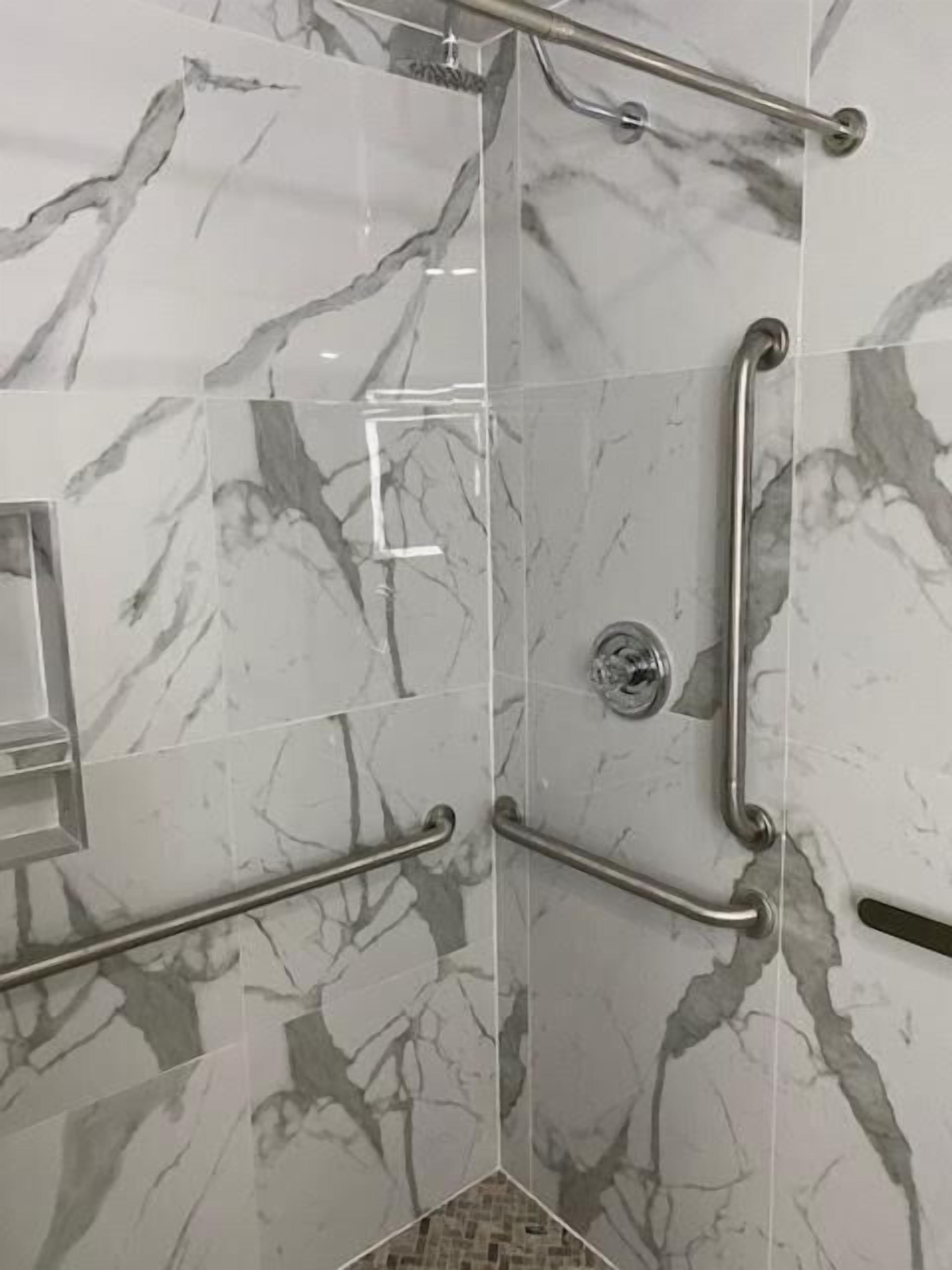 Un baño con baldosas de mármol, ducha con cabezal de ducha manual y barra de apoyo.