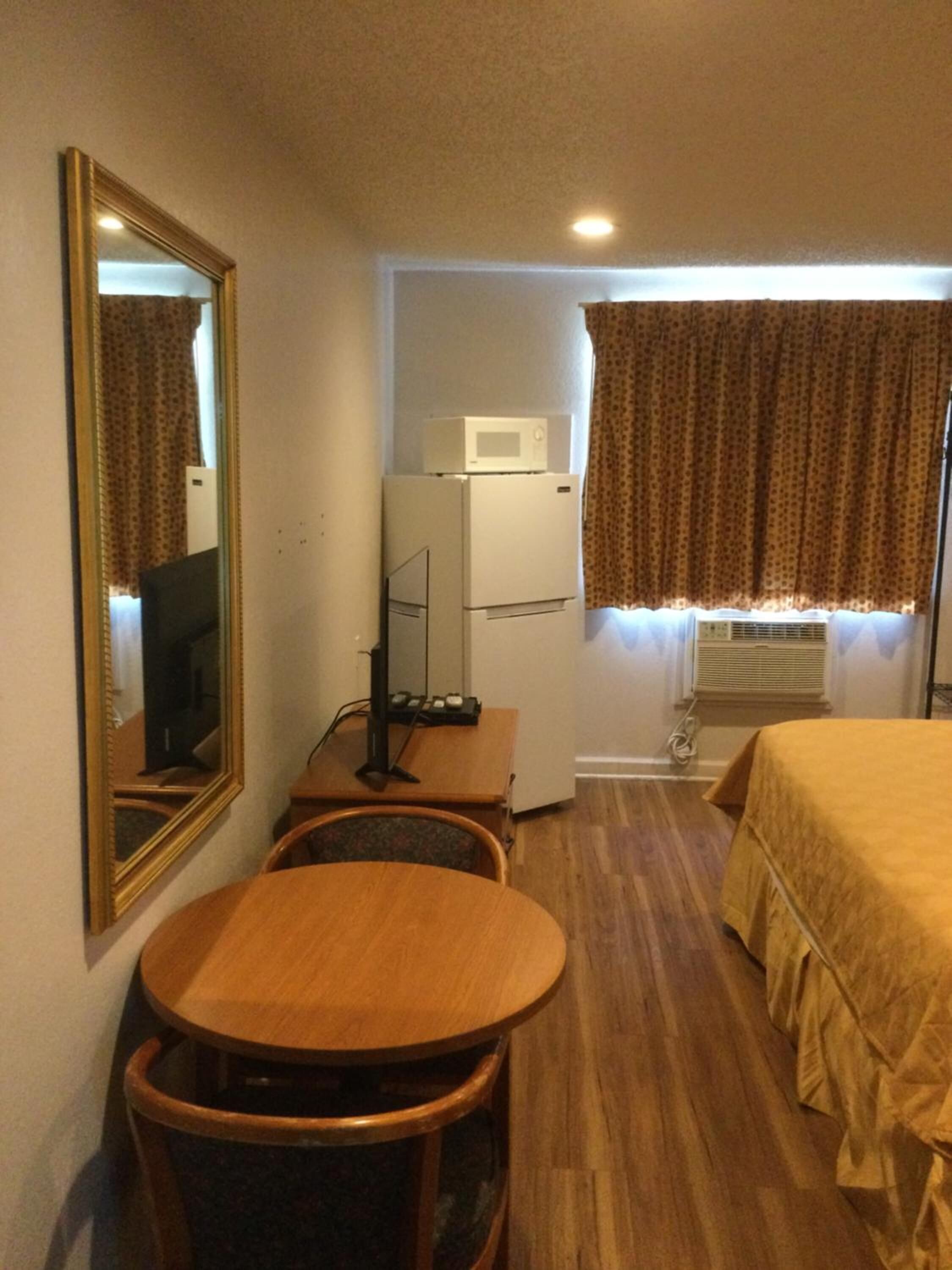 Habitación de hotel con cama, escritorio con computadora, refrigerador pequeño, microondas, ventana con cortinas y espejo en la pared.
