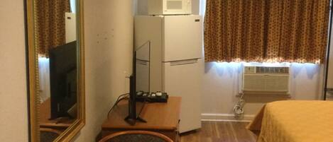Habitación de hotel con cama, escritorio con computadora, refrigerador pequeño, microondas, ventana con cortinas y espejo en la pared.