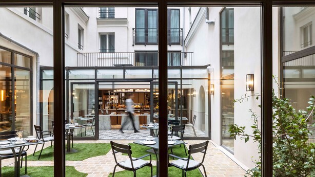 A courtyard with tables and chairs, a glass wall, and a view of a restaurant inside.