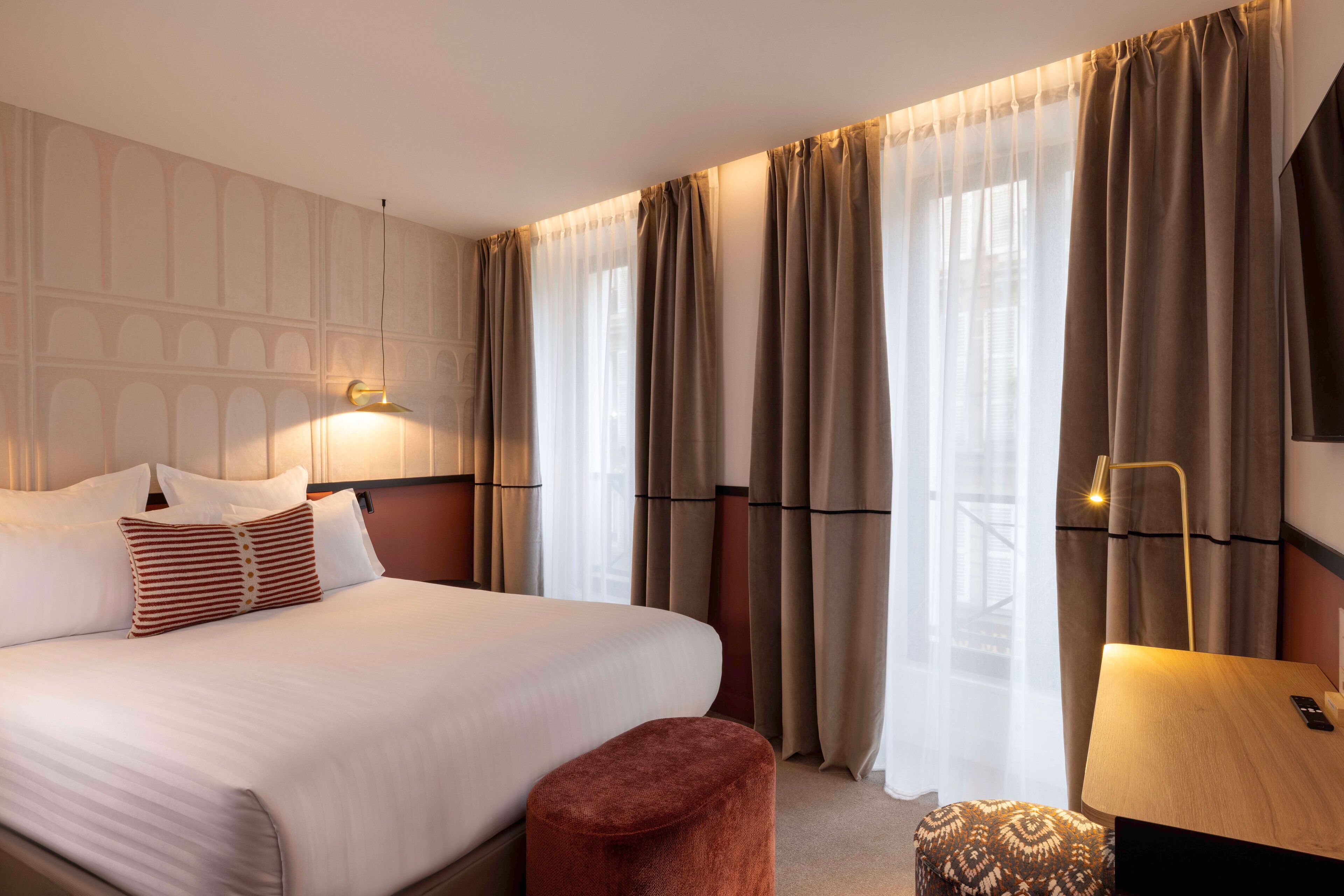 A hotel room with a large bed, a bedside table with a lamp, and a view through sheer curtains.
