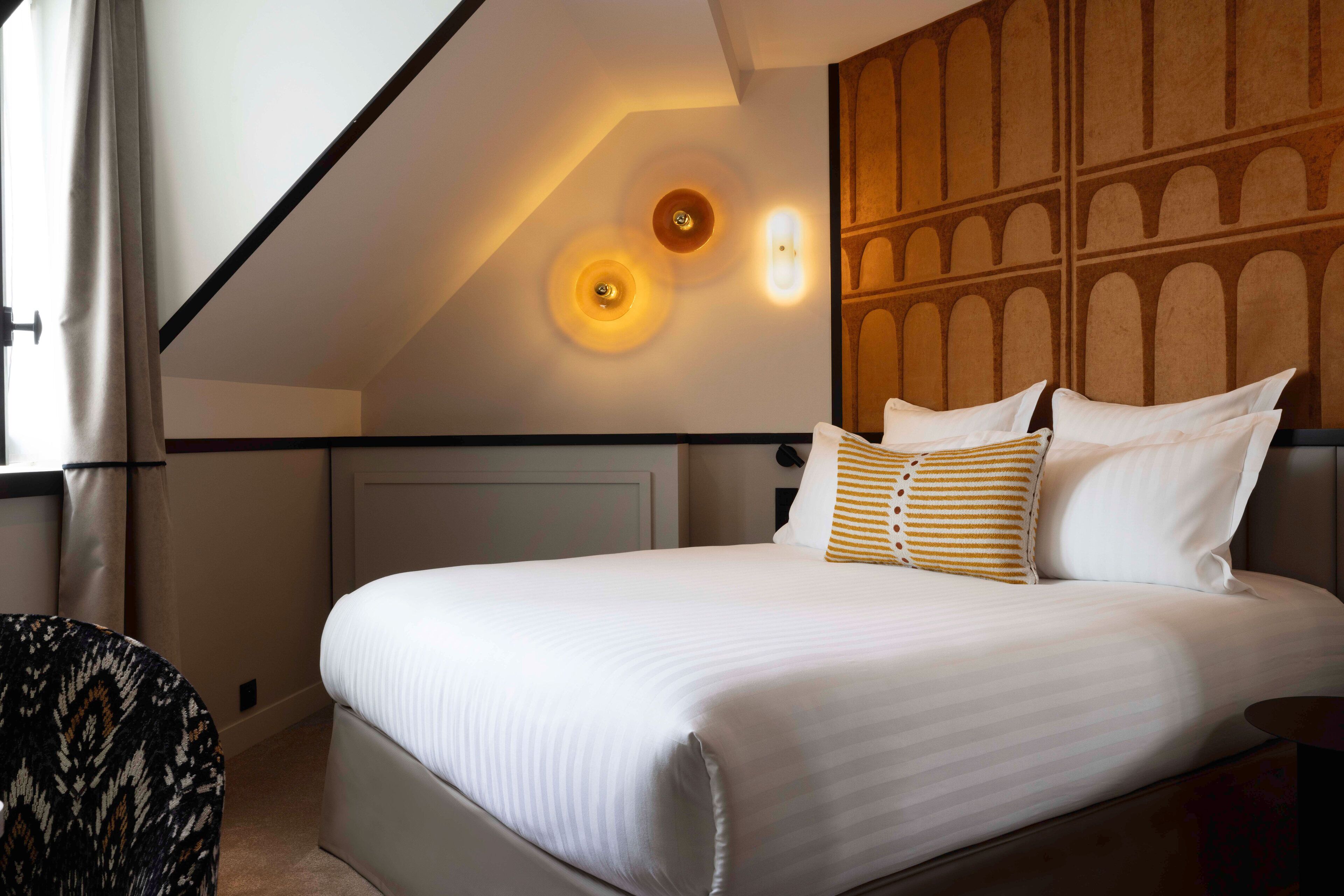 A neatly made bed with white linens and striped pillows, a wooden headboard, and a wall with a geometric pattern.