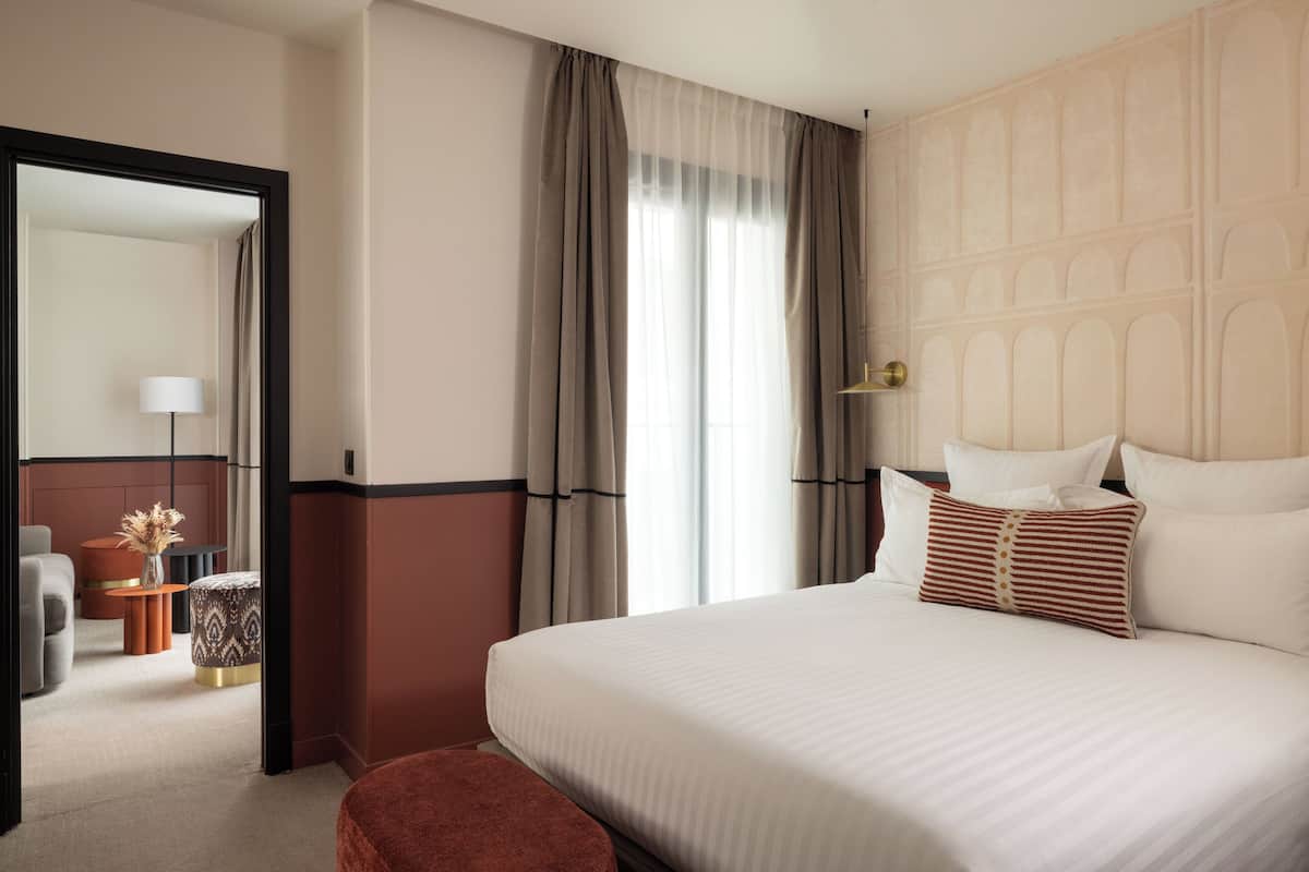 A hotel room with a large bed, a headboard with a geometric pattern, a small round table with a vase, and a mirror in the corner.