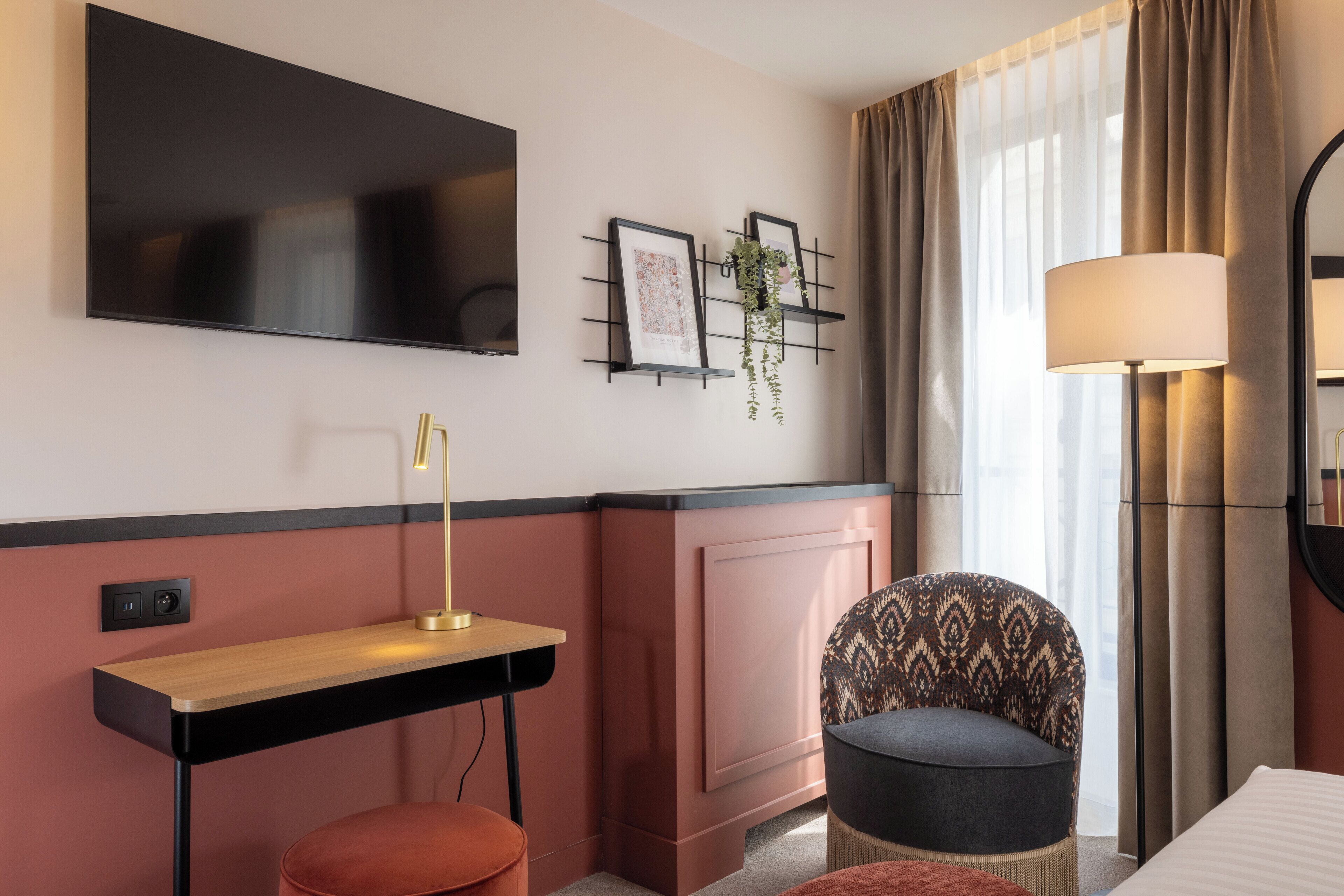 Una habitación de hotel moderna con televisión de pantalla plana, un escritorio con lámpara, una silla estampada y un ventanal con cortinas.