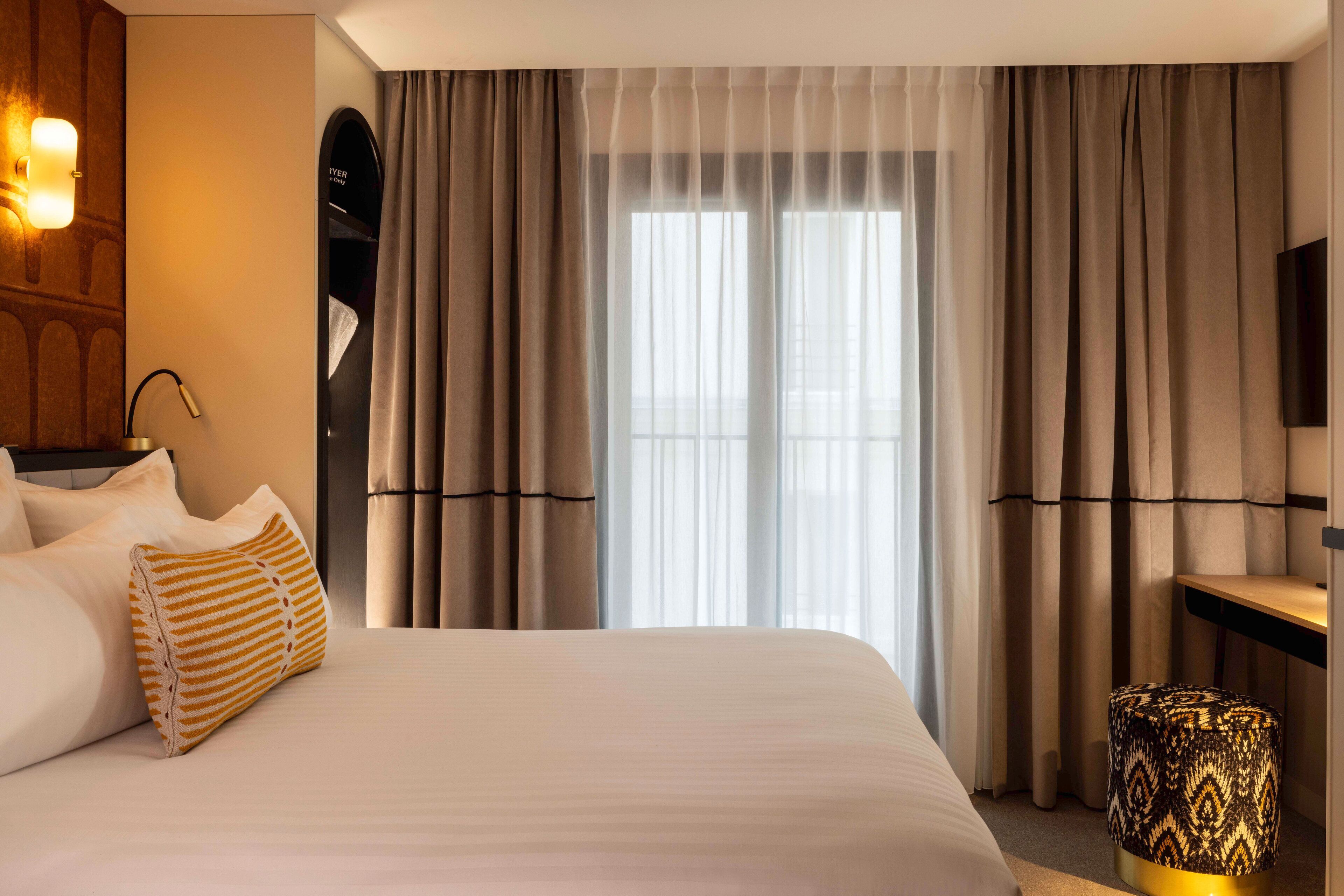 A hotel room with a large bed, a bedside table, a lamp, and a window with curtains.