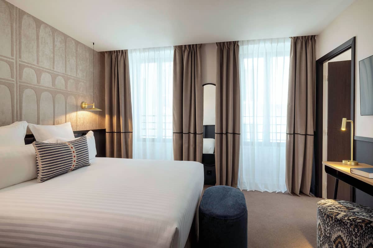 A hotel room with a large bed, a bedside table with a lamp, a mirror, and a window with curtains.