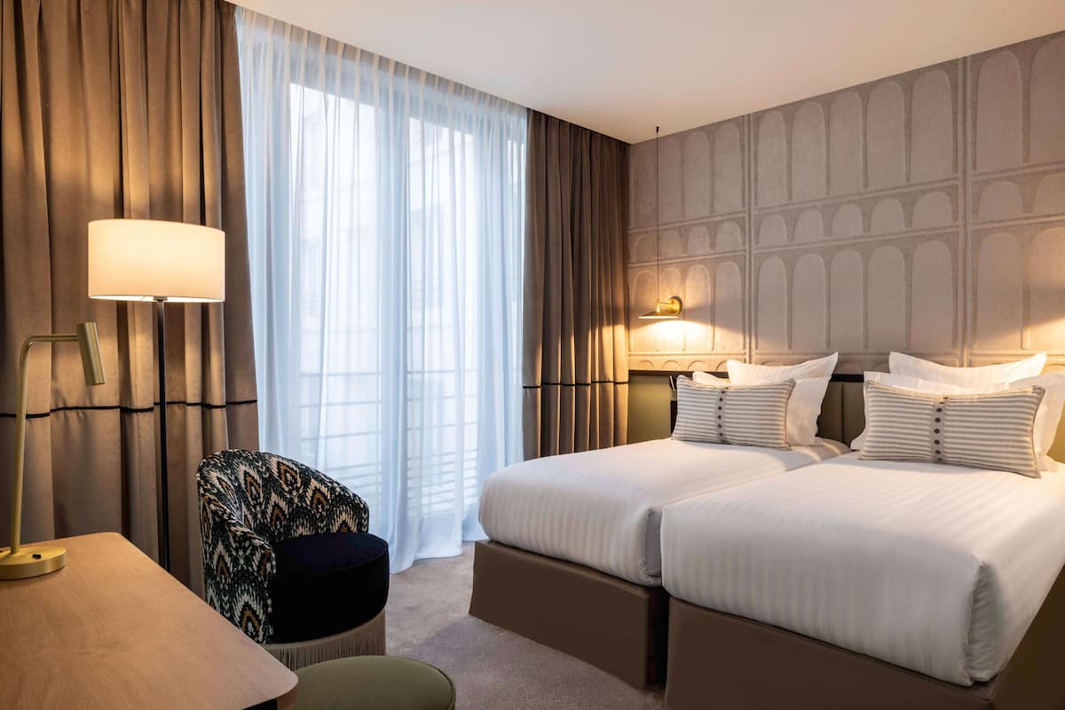 A hotel room with two beds, a desk, a chair, and a large window with curtains.