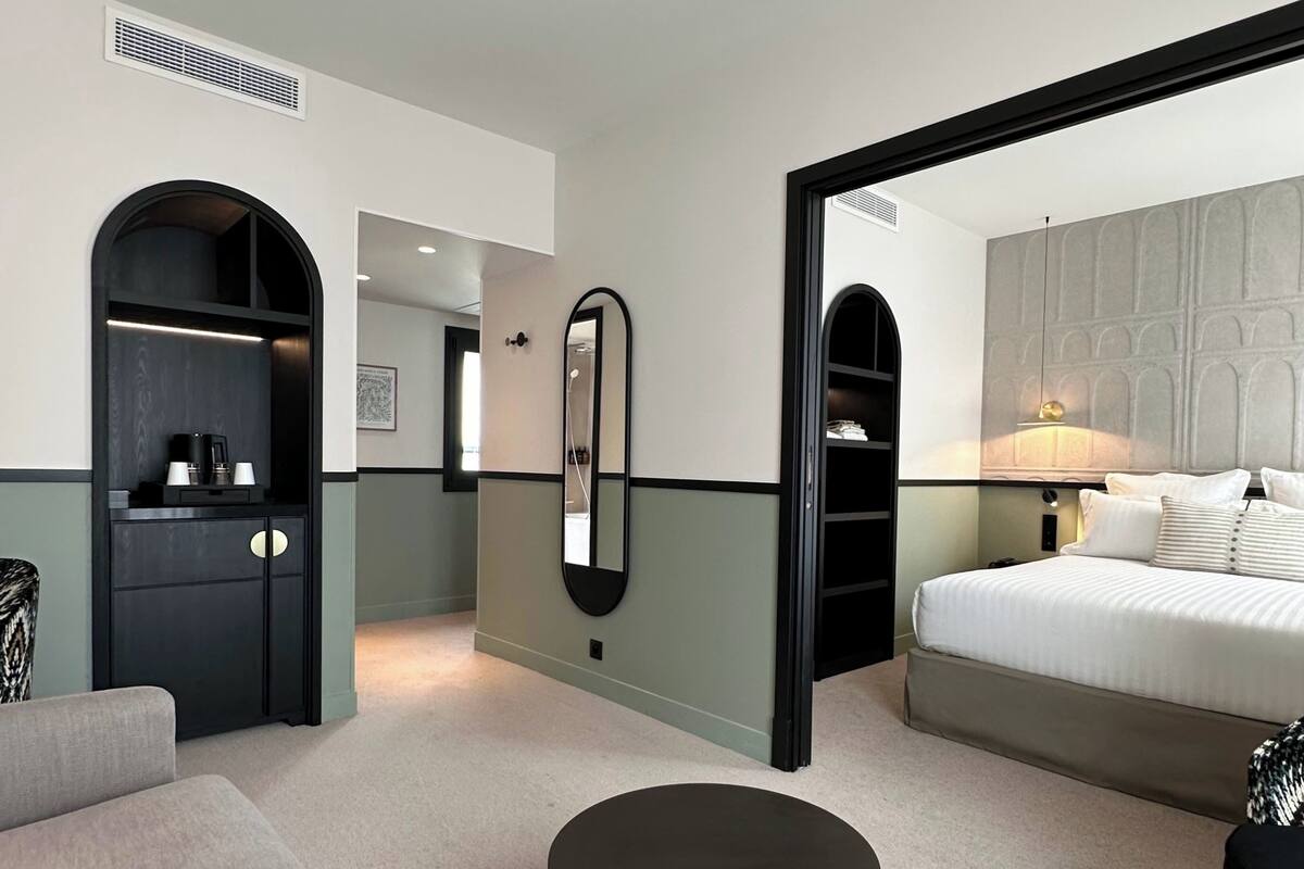 A modern hotel room with a large bed, a round black ottoman, a wardrobe with a mirror, and a seating area with a sofa.
