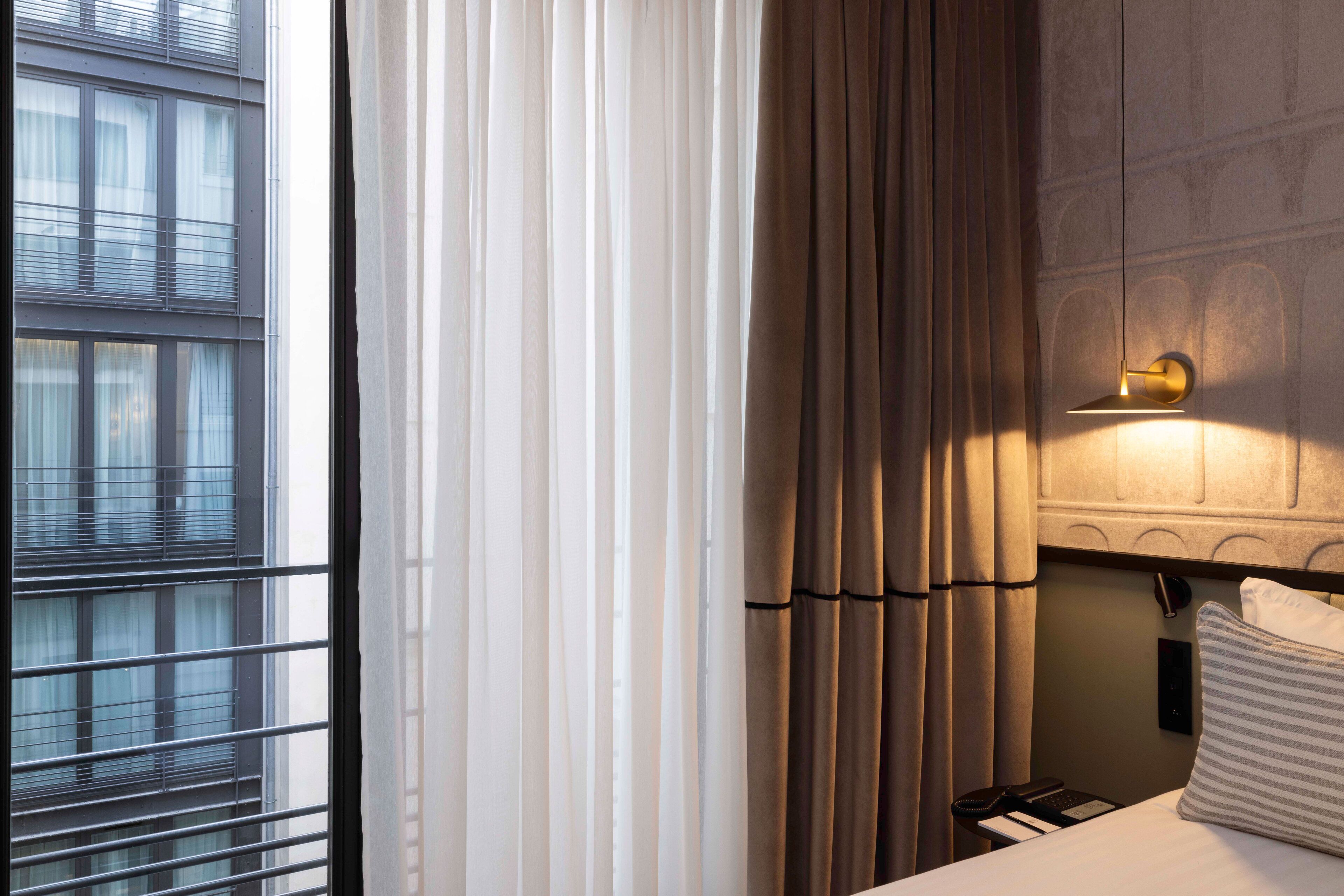 A hotel room with a large window, curtains, a bedside lamp, a bed with a pillow, and a nightstand with a phone and remote control.