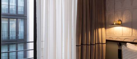 A hotel room with a large window, curtains, a bedside lamp, a bed with a pillow, and a nightstand with a phone and remote control.