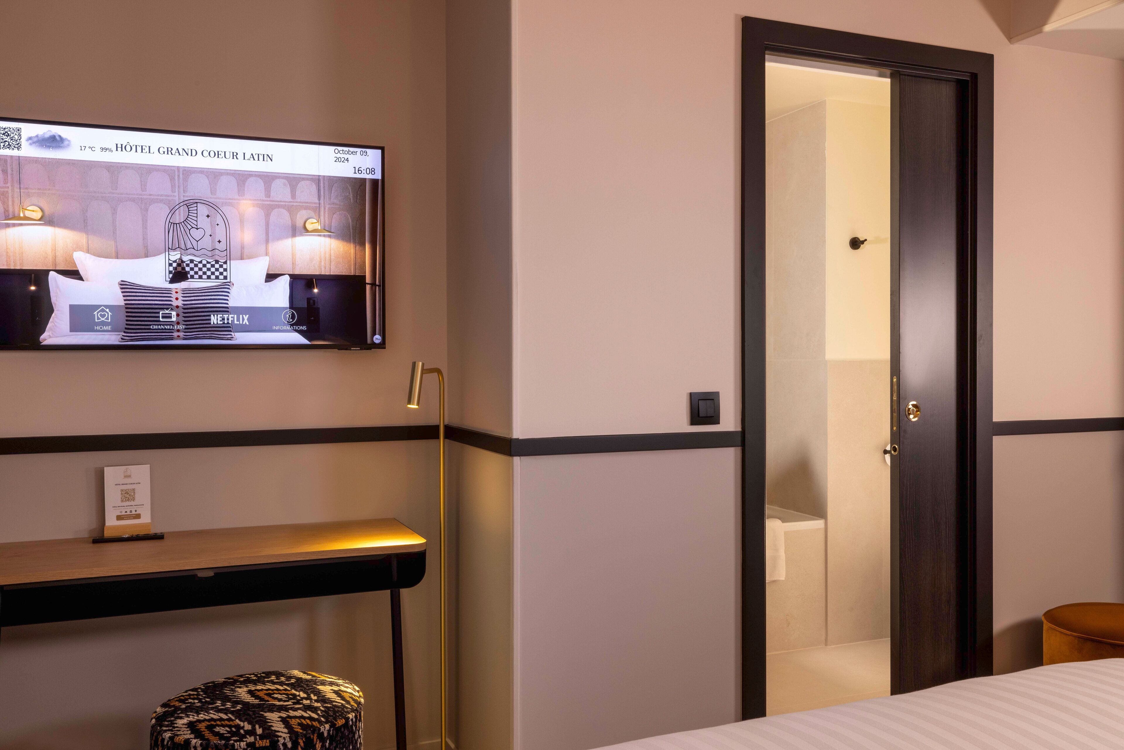 Habitació d'hotel amb un llit, una taula de nit, una televisió que mostra el nom de l'hotel i una porta que dona a un bany.