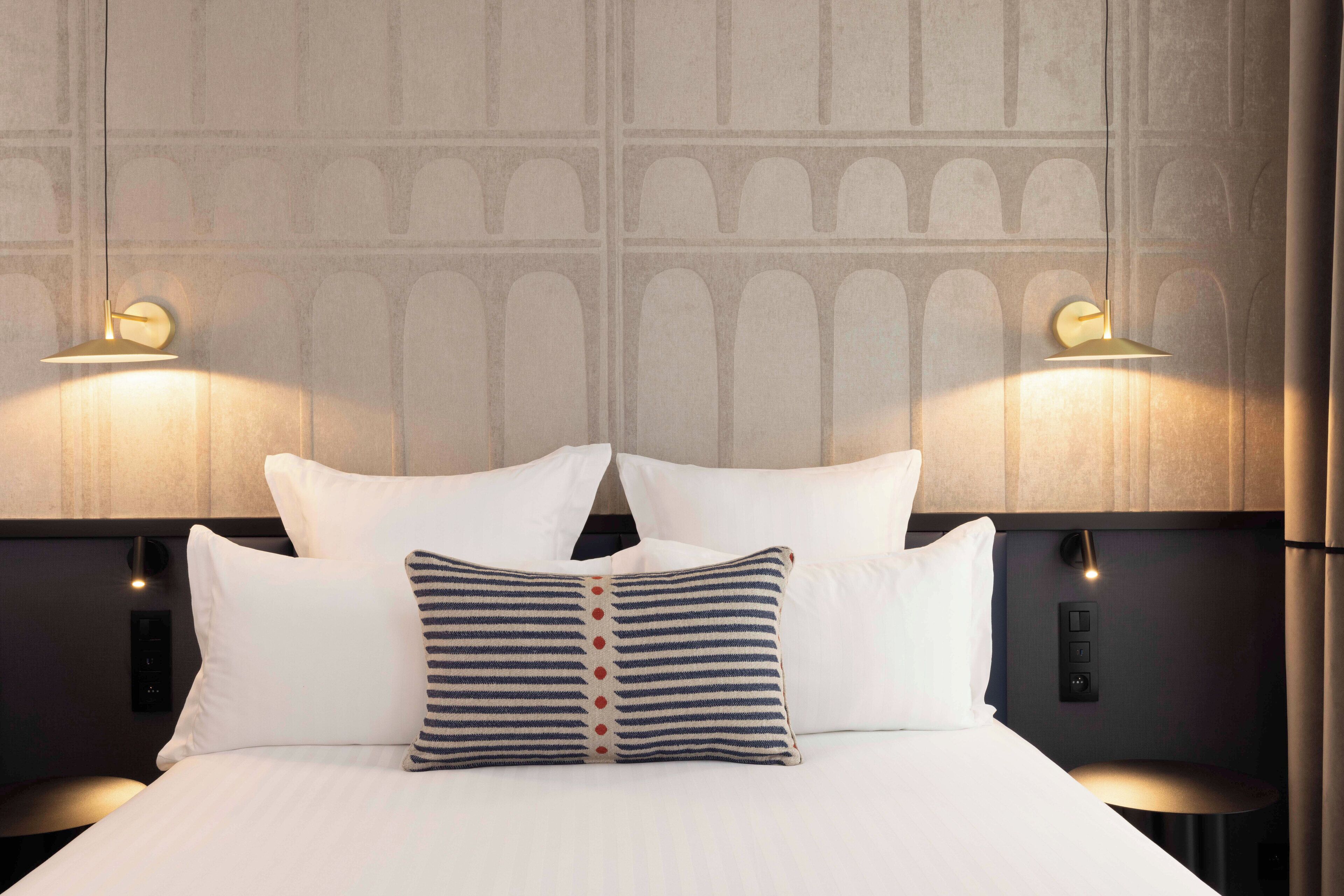 Una cama bien hecha con sábanas blancas y almohadas a rayas, dos lámparas fijadas a la pared y un diseño de pared con textura.