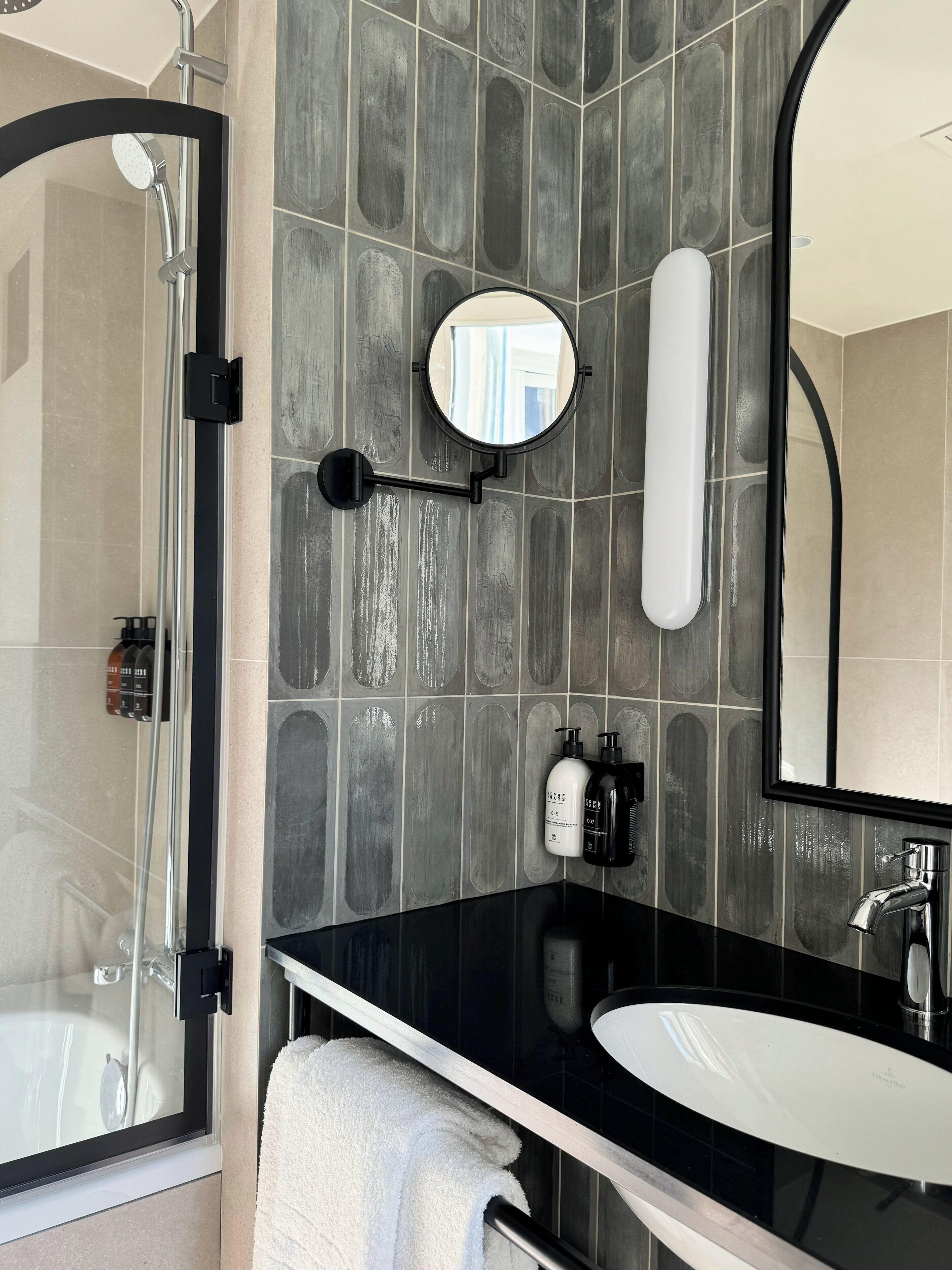 A modern bathroom with a glass shower enclosure, a black countertop, a white sink, and a large mirror.