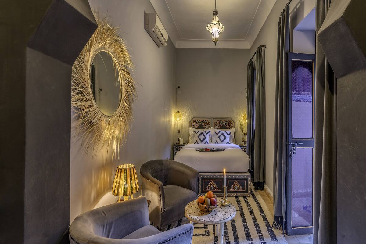 A modern bedroom with a bed, two armchairs, a small table with fruit, a decorative mirror, and a chandelier.